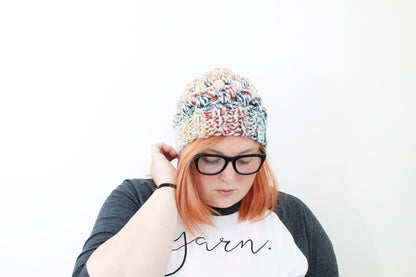 Chunky rainbow color puff stitch beanie on model with peach hair and thick black glasses